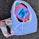 A light blue fabric pouch with a white drawstring closure, laid on asphalt next to a white tray holding sunglasses, a scrunchie, and a book titled 'Person in progress'. The pouch has a small white tag reading 'ilora'.