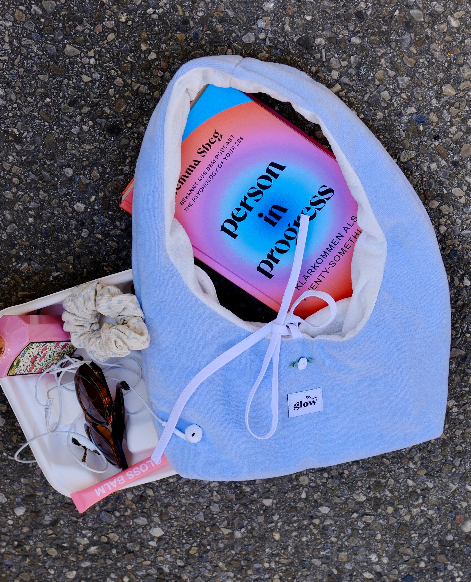 A light blue fabric pouch with a white drawstring closure, laid on asphalt next to a white tray holding sunglasses, a scrunchie, and a book titled 'Person in progress'. The pouch has a small white tag reading 'ilora'.