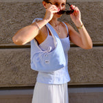 A woman wearing a light blue ribbed camisole top and a long white skirt, carrying a light blue shoulder bag with a small white label, standing against a textured brown wall.
