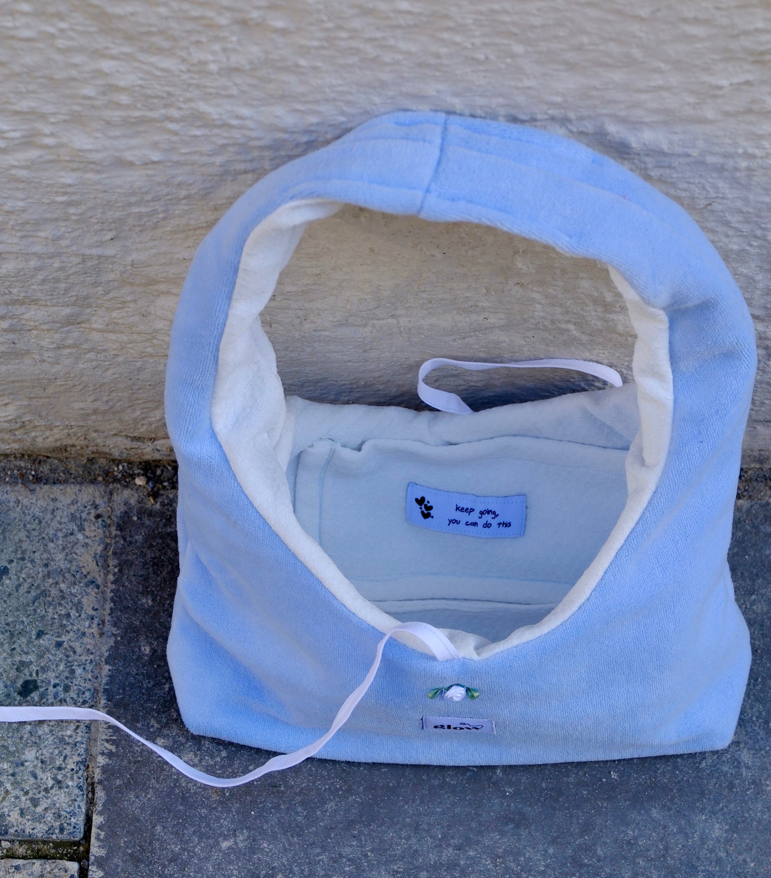 A light blue fleece shoulder bag with a white interior lining, placed on a gray stone surface against a beige stucco wall. The brand name 'KONG PET' is visible on a small blue label inside the bed.