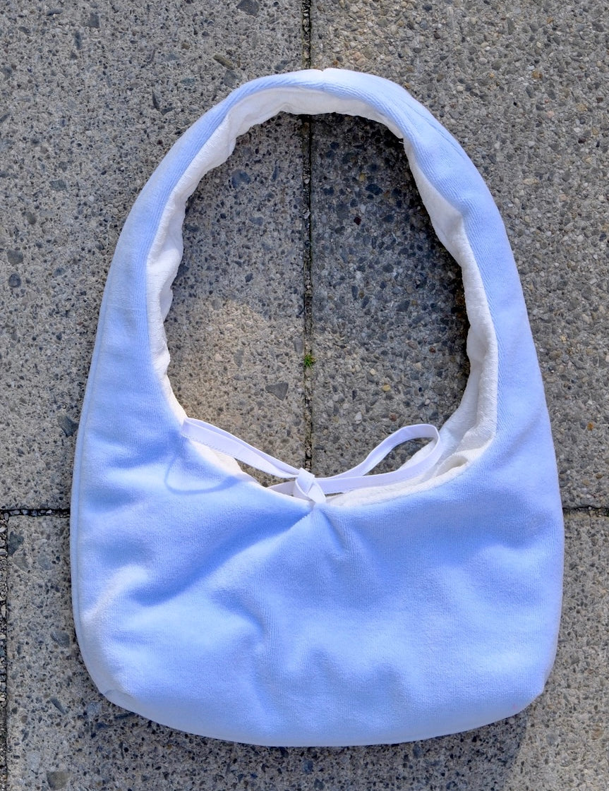 A light blue fabric shoulder bag with a white lining and white tie detail, laid flat on gray concrete pavers.