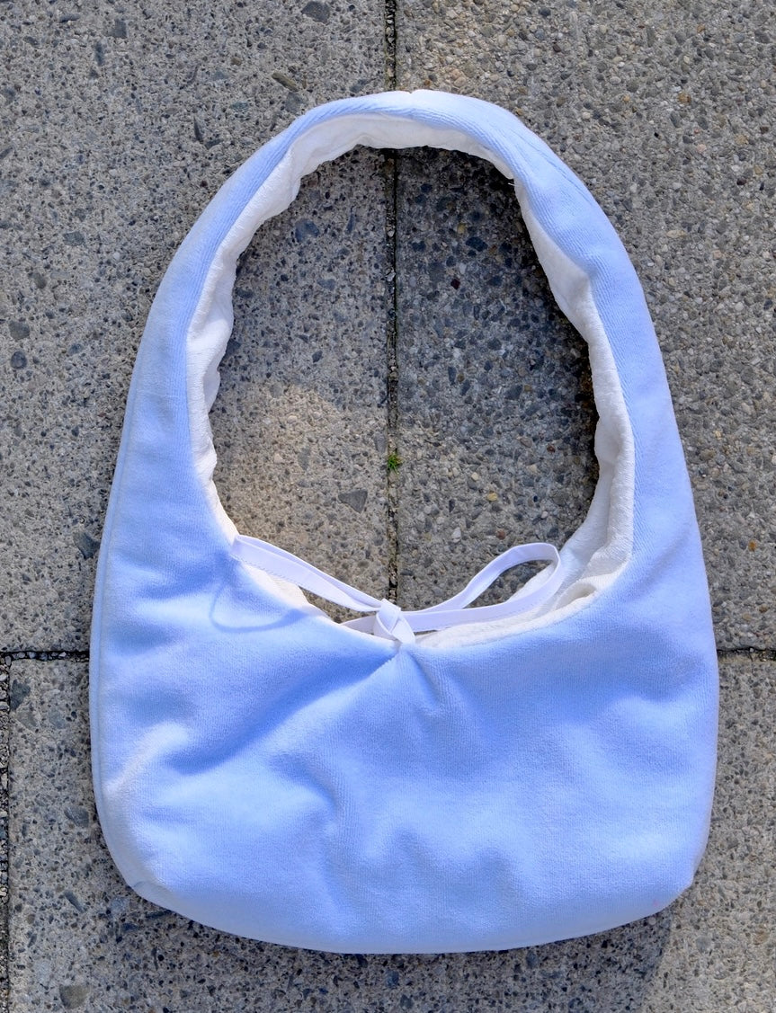A light blue fabric shoulder bag with a white lining and white tie detail, laid flat on gray concrete pavers.