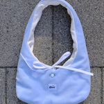 A light blue fabric tote bag with a white ribbon tie and a small white button, laid flat on gray concrete pavers. A small label on the front reads 'glow'.