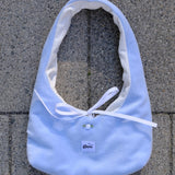 A light blue fabric tote bag with a white ribbon tie and a small white button, laid flat on gray concrete pavers. A small label on the front reads 'glow'.