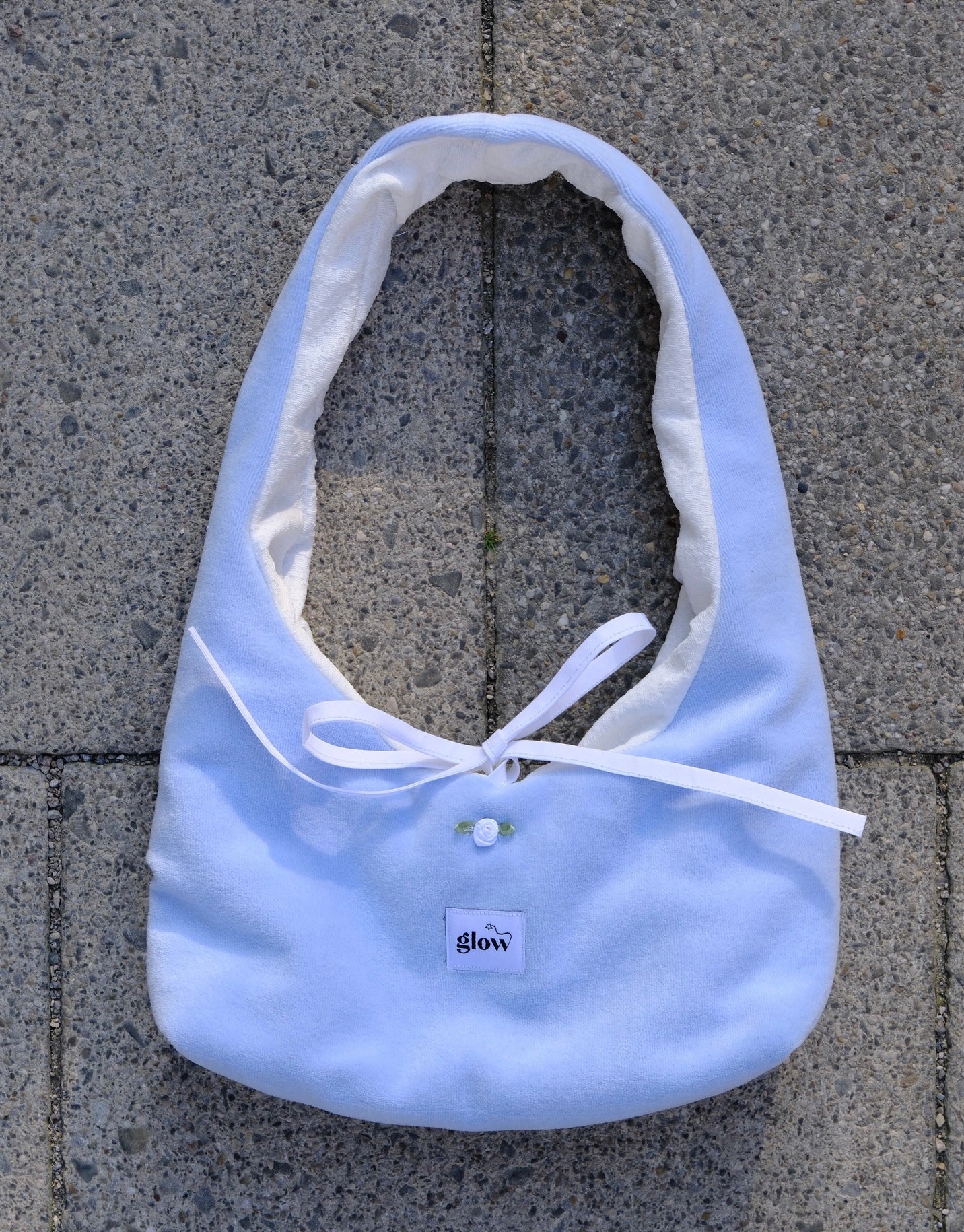 A light blue fabric tote bag with a white ribbon tie and a small white button, laid flat on gray concrete pavers. A small label on the front reads 'glow'.