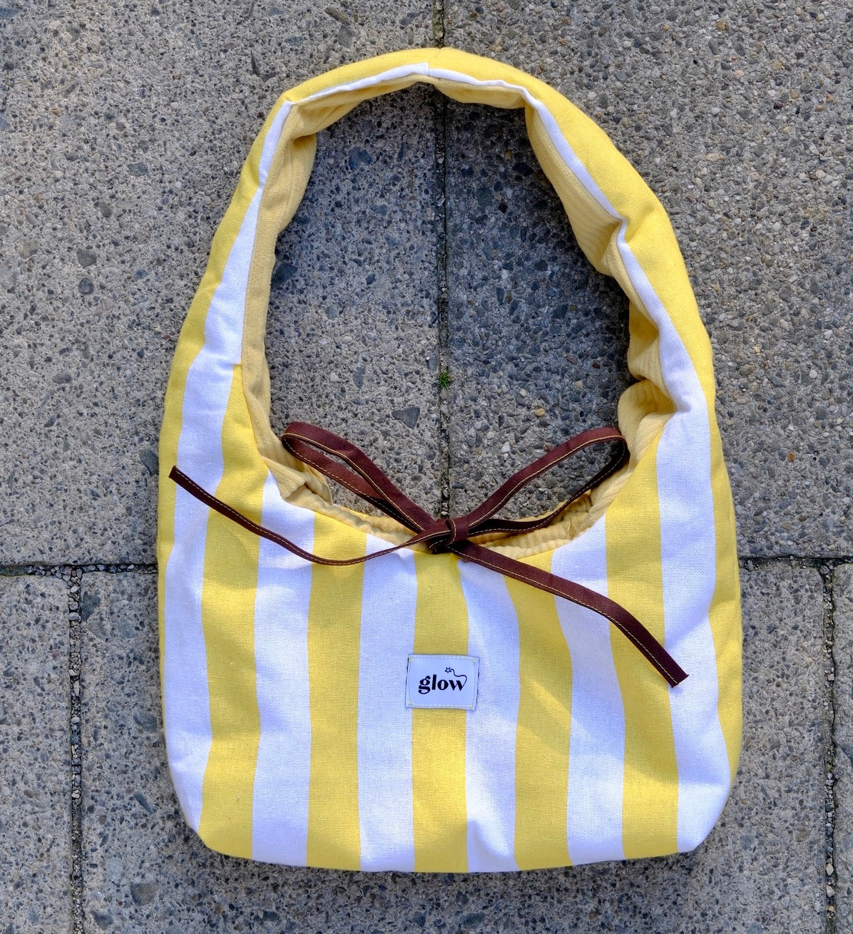 A yellow and white striped fabric bag with a dark brown ribbon tied in a bow, laid flat on gray concrete pavers. A small white label on the front reads 'glow'.
