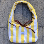 A yellow and white striped fabric bag with a dark brown ribbon tied in a bow, laid flat on gray concrete pavers. A small white label on the front reads 'glow'.