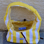 A handmade fabric shoulder bag with yellow and white vertical stripes, featuring a yellow corduroy interior lining and a dark brown leather strap, set against a textured beige wall.