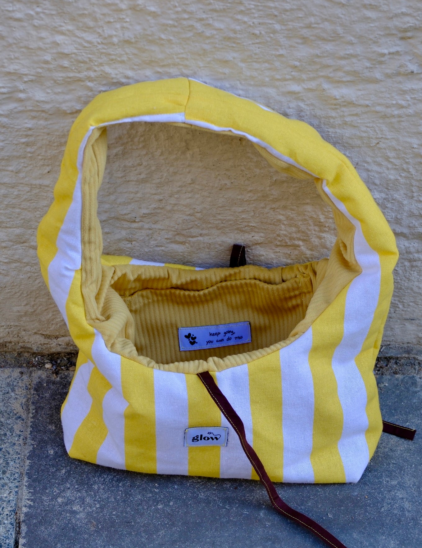 A handmade fabric shoulder bag with yellow and white vertical stripes, featuring a yellow corduroy interior lining and a dark brown leather strap, set against a textured beige wall.