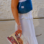 A woman wearing a white tiered maxi dress and a blue denim vest-style bag, holding a magazine in front of a textured white wall.