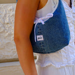 A person wearing a white sleeveless top and white skirt carries a blue denim shoulder bag with a white drawstring bow and a small white brand label that reads 'glow'.