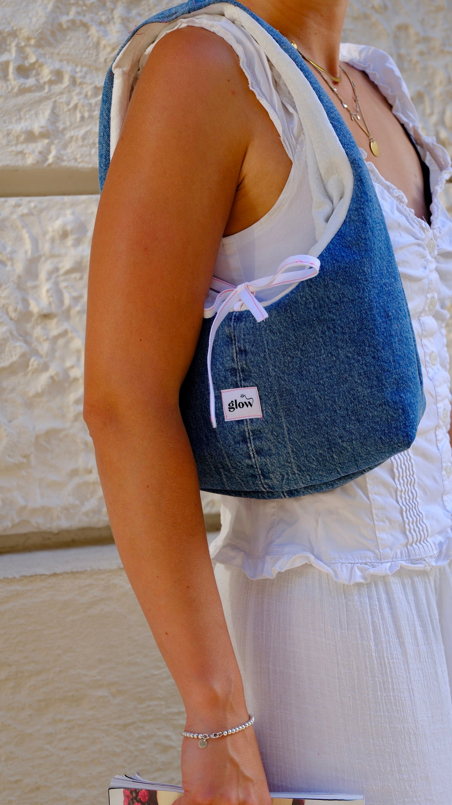 A person wearing a white sleeveless top and white skirt carries a blue denim shoulder bag with a white drawstring bow and a small white brand label that reads 'glow'.