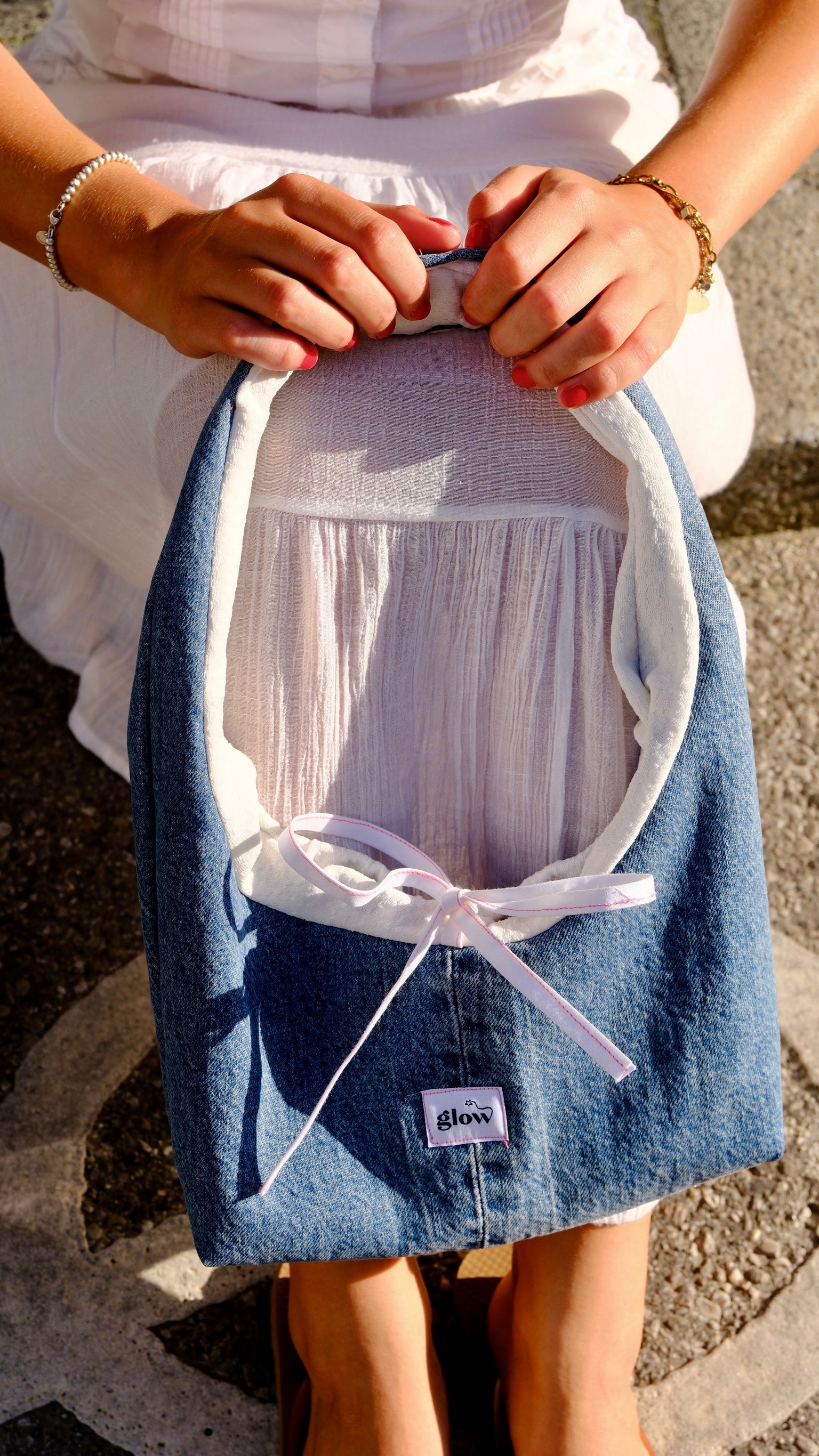 A person holds a denim shoulder bag with a white inner lining and a white ribbon bow. The pouch has a small white label that reads 'glow'.