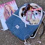 A blue denim tote bag laid on asphalt with a white zippered pocket and a pink ribbon bow, surrounded by everyday accessories including sunglasses, a pink camera, earphones, a magazine, and a scrunchie.