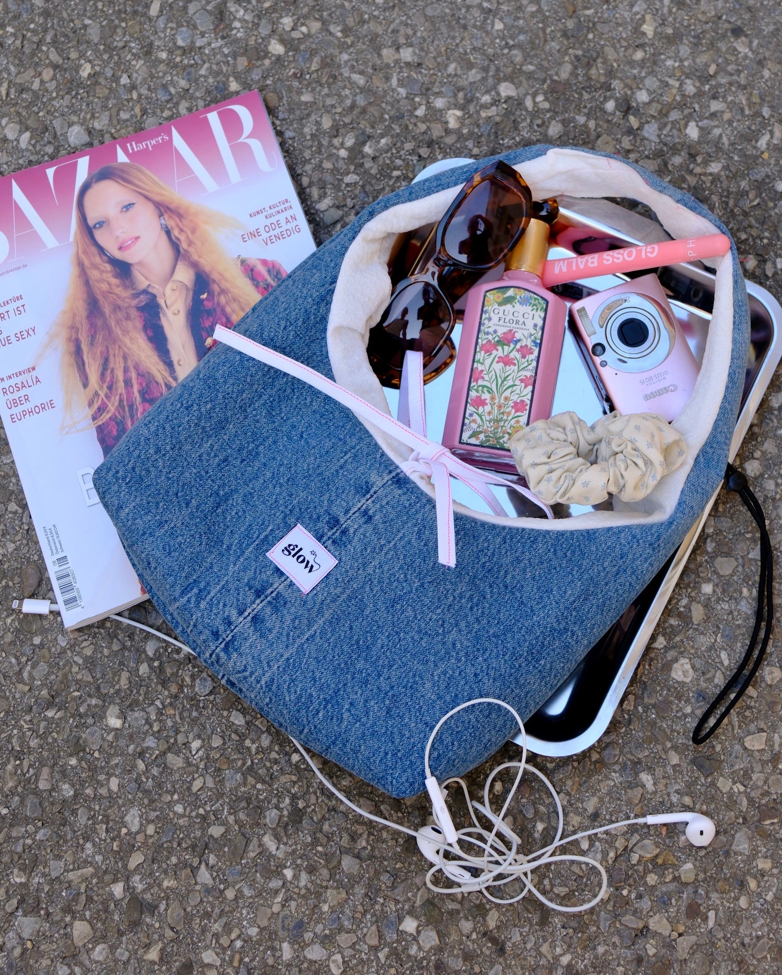 A blue denim tote bag laid on asphalt with a white zippered pocket and a pink ribbon bow, surrounded by everyday accessories including sunglasses, a pink camera, earphones, a magazine, and a scrunchie.