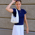 A woman wearing a navy blue t-shirt and a long white skirt stands against a textured beige wall, carrying a white patterned tote bag over her shoulder.