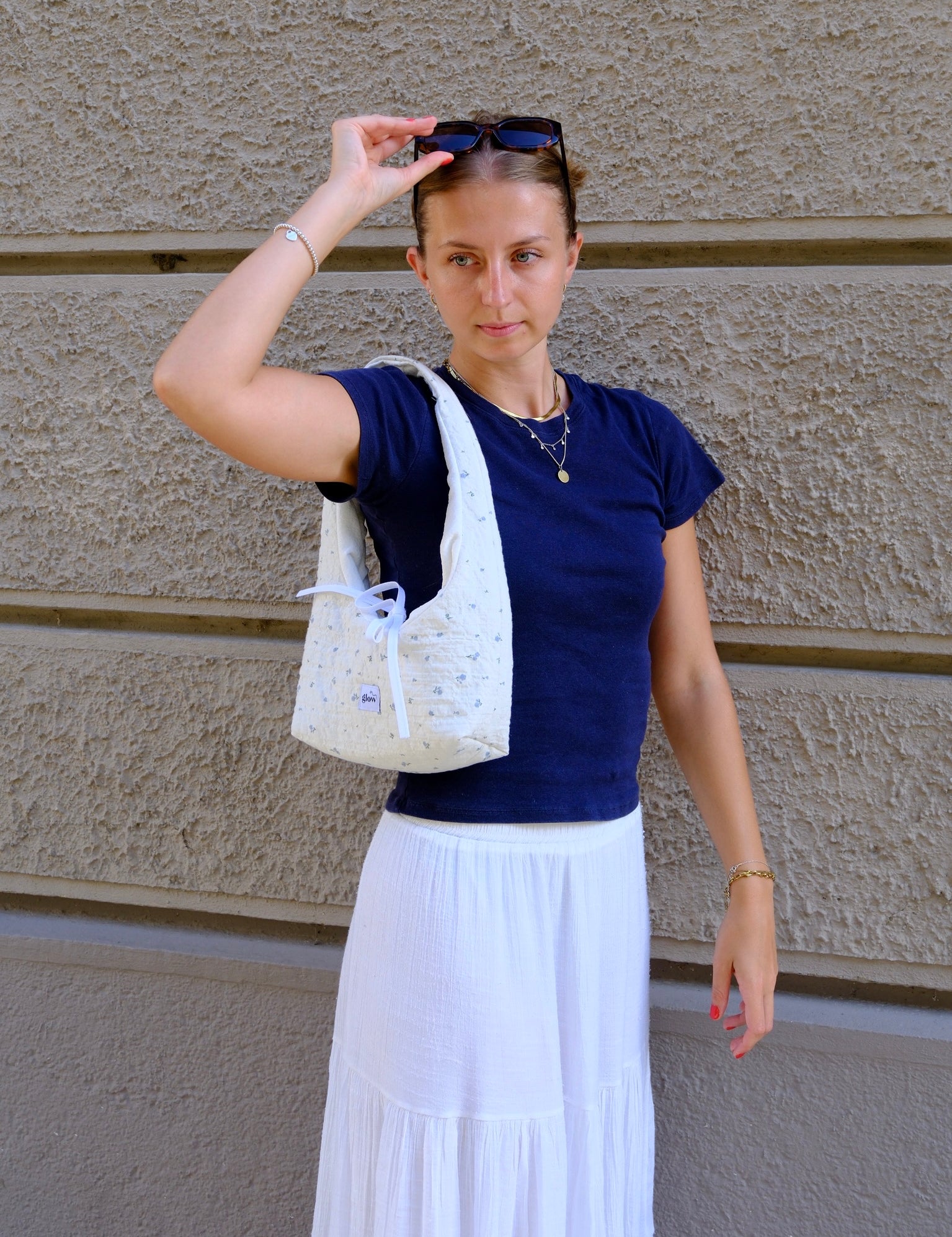 A woman wearing a navy blue t-shirt and a long white skirt stands against a textured beige wall, carrying a white patterned tote bag over her shoulder.