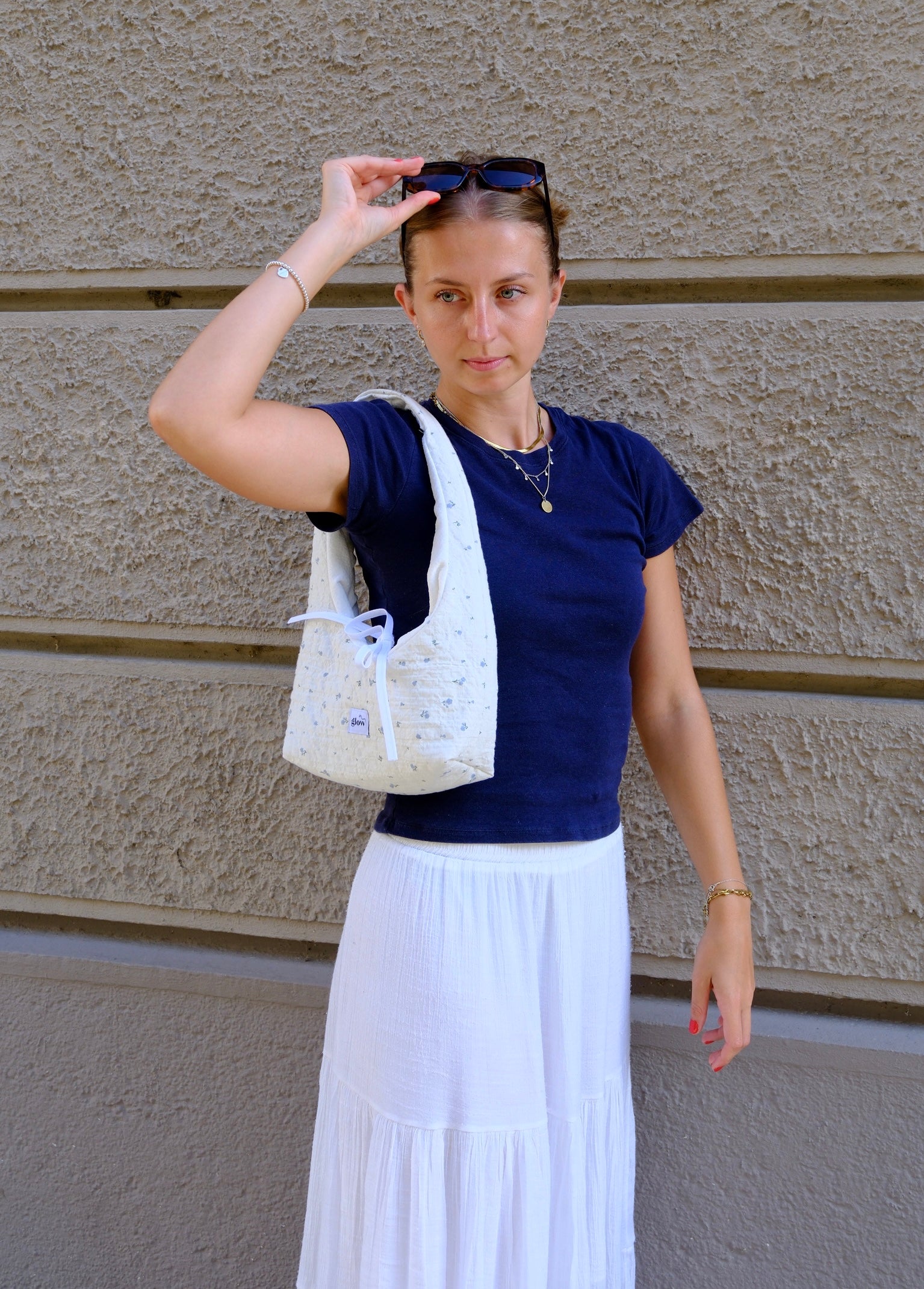 A woman wearing a navy blue t-shirt and a long white skirt stands against a textured beige wall, carrying a white patterned tote bag over her shoulder.