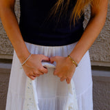 A person wearing a black t-shirt and a long white skirt holds a cream-colored quilted handbag with a white ribbon tie and small blue floral pattern. The background shows a textured beige wall.