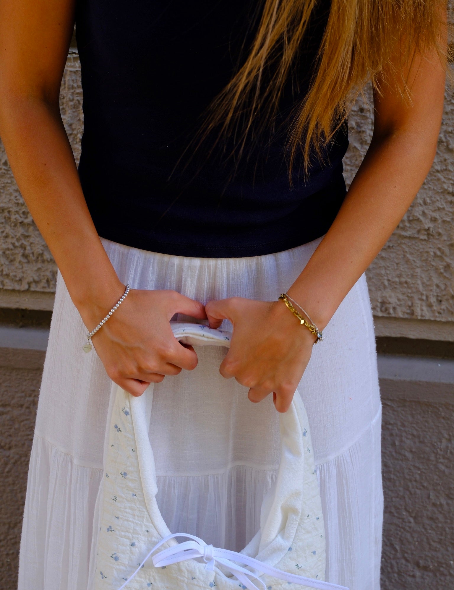 A person wearing a black t-shirt and a long white skirt holds a cream-colored quilted handbag with a white ribbon tie and small blue floral pattern. The background shows a textured beige wall.