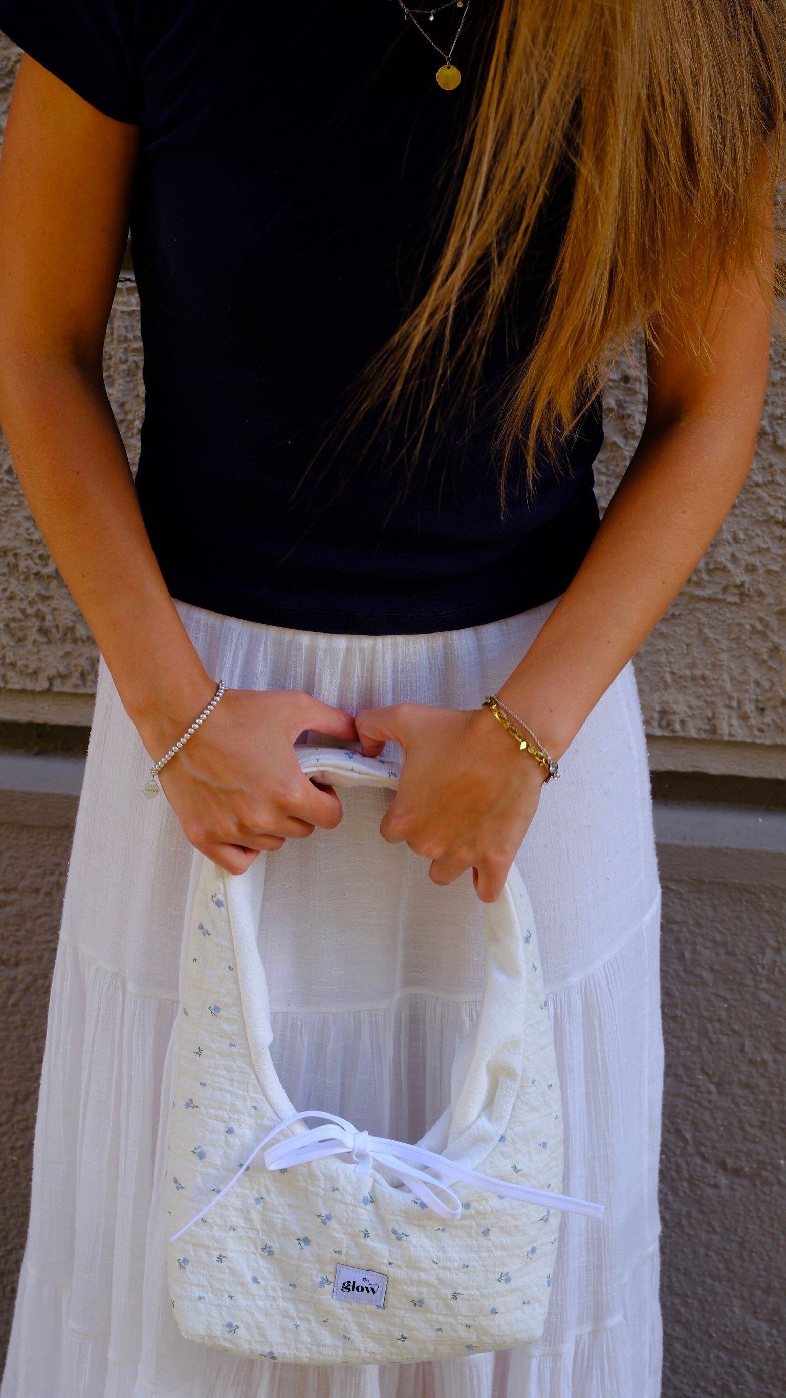 A person wearing a black t-shirt and a long white skirt holds a cream-colored quilted handbag with a white ribbon tie and small blue floral pattern. The background shows a textured beige wall.