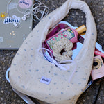A cream-colored fabric shoulder bag with a small blue floral pattern, styled on a silver tray with various accessories like a pink bottle, a scrunchie, and earphones on an asphalt surface.