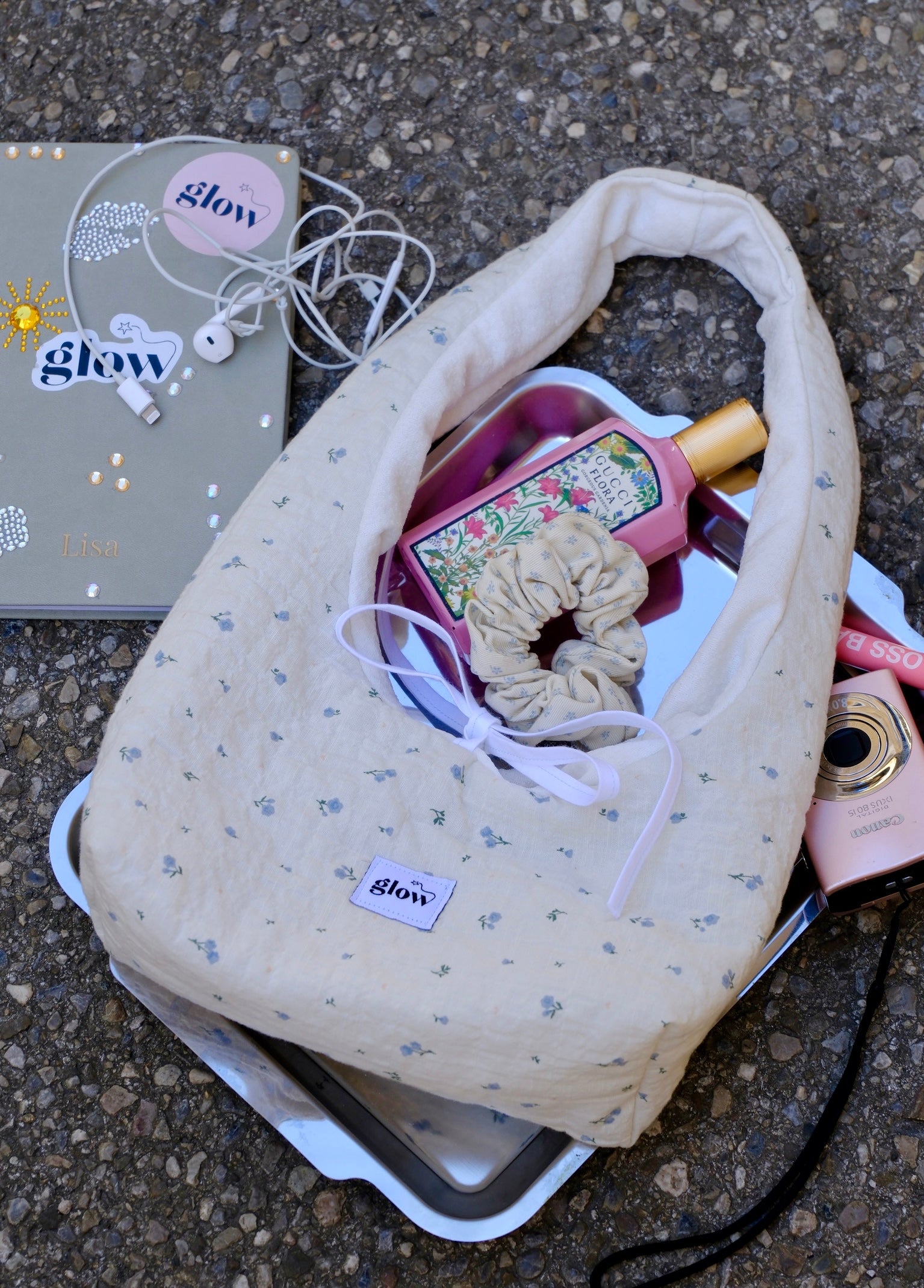 A cream-colored fabric shoulder bag with a small blue floral pattern, styled on a silver tray with various accessories like a pink bottle, a scrunchie, and earphones on an asphalt surface.