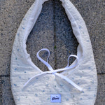 A white quilted baby bib with a tiny blue floral pattern and white ribbon ties, laid flat on gray concrete pavement. A small square brand label reading 'glow' is visible near the bottom.