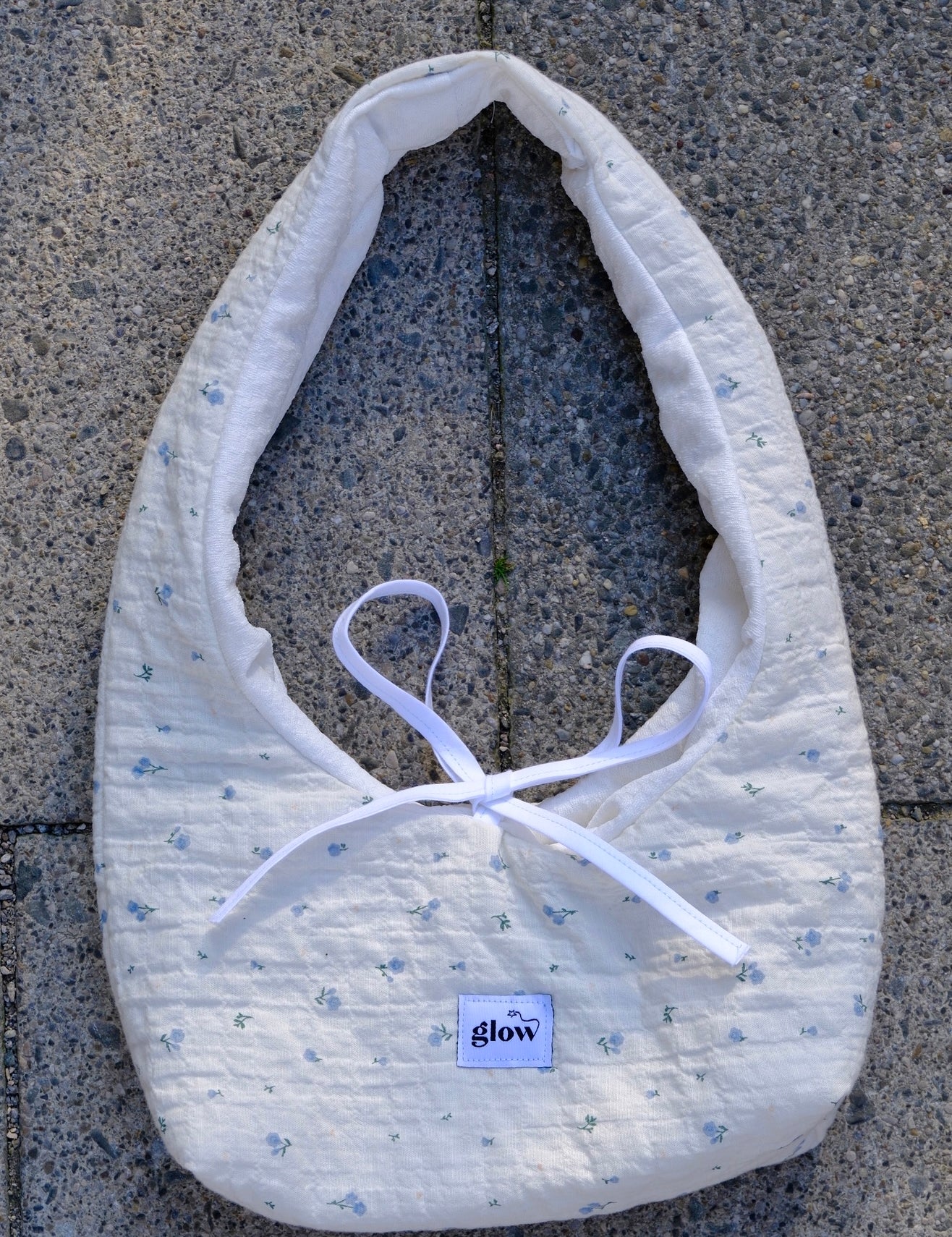 A white quilted baby bib with a tiny blue floral pattern and white ribbon ties, laid flat on gray concrete pavement. A small square brand label reading 'glow' is visible near the bottom.