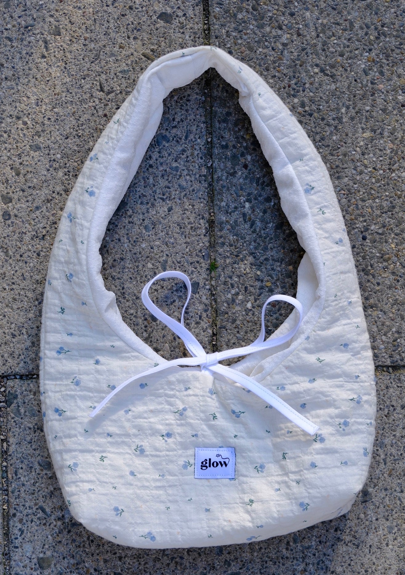 A white quilted baby bib with a tiny blue floral pattern and white ribbon ties, laid flat on gray concrete pavement. A small square brand label reading 'glow' is visible near the bottom.