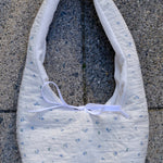 A white quilted fabric shoulder bag with a tiny blue floral pattern and a white ribbon bow, laid flat on gray concrete pavement.