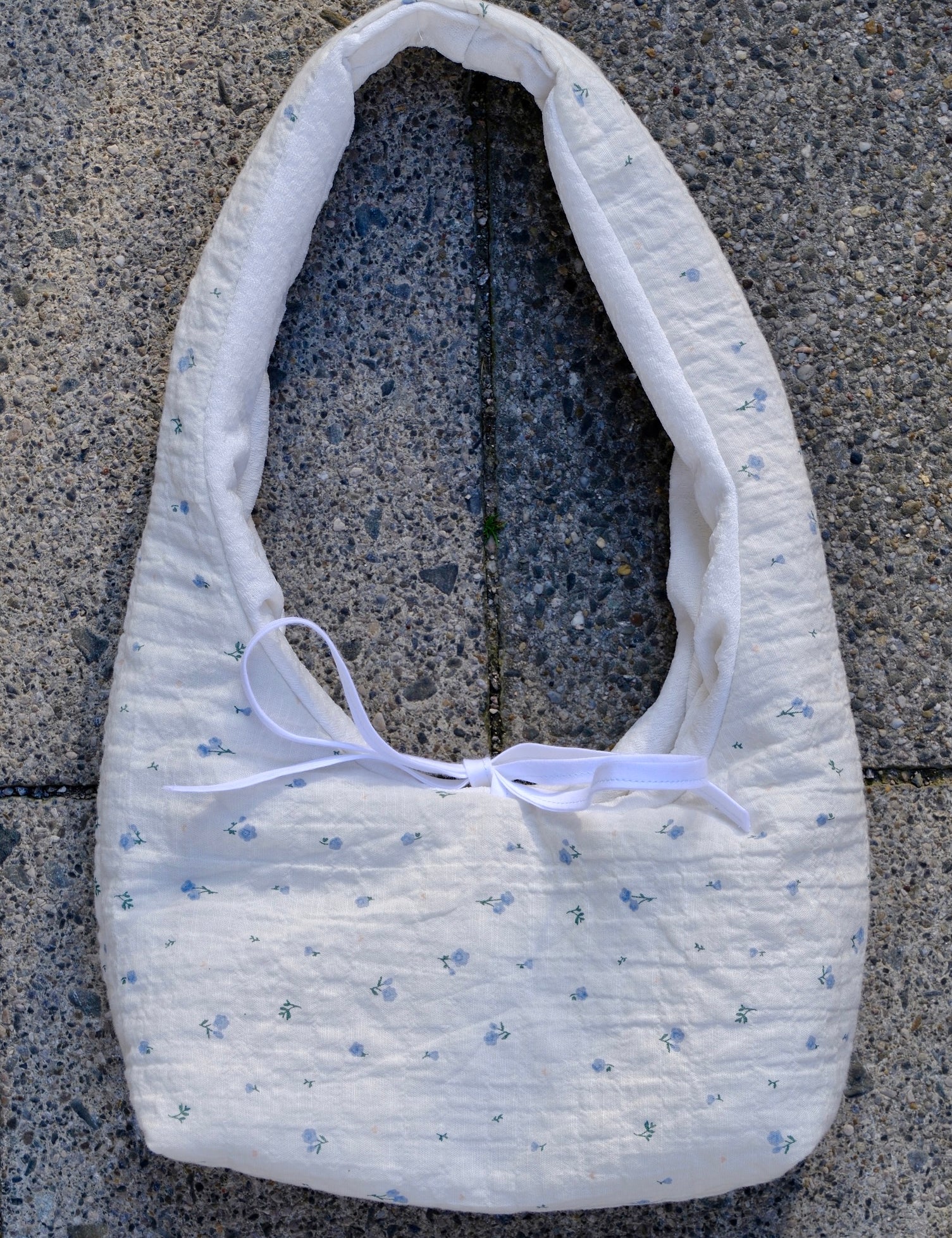 A white quilted fabric shoulder bag with a tiny blue floral pattern and a white ribbon bow, laid flat on gray concrete pavement.