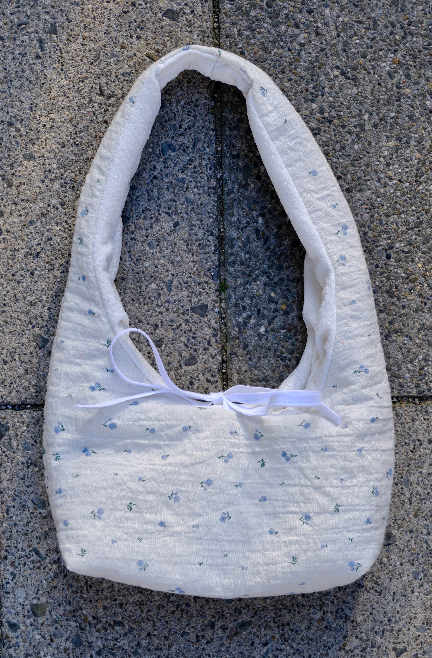 A white quilted fabric shoulder bag with a tiny blue floral pattern and a white ribbon bow, laid flat on gray concrete pavement.