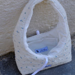 A handmade quilted fabric handbag with a white base, small blue floral print, and a white strap, resting on a gray stone surface against a beige stucco wall.