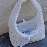 A handmade quilted fabric handbag with a white base, small blue floral print, and a white strap, resting on a gray stone surface against a beige stucco wall.