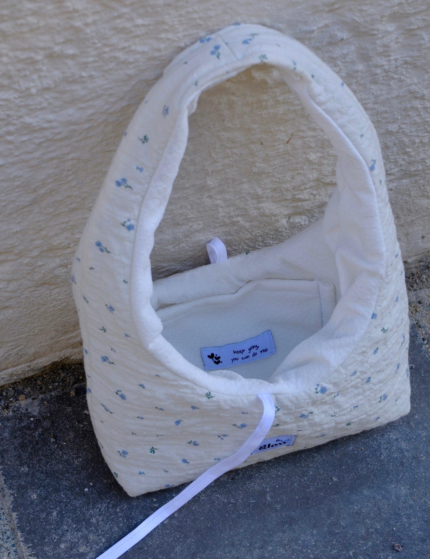 A handmade quilted fabric handbag with a white base, small blue floral print, and a white strap, resting on a gray stone surface against a beige stucco wall.