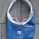 A blue denim shoulder bag with a white interior lining and a light purple tie, laid flat on gray concrete pavement. A small white label on the front reads 'glow'.