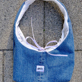 A blue denim shoulder bag with a white interior lining and a light purple tie, laid flat on gray concrete pavement. A small white label on the front reads 'glow'.