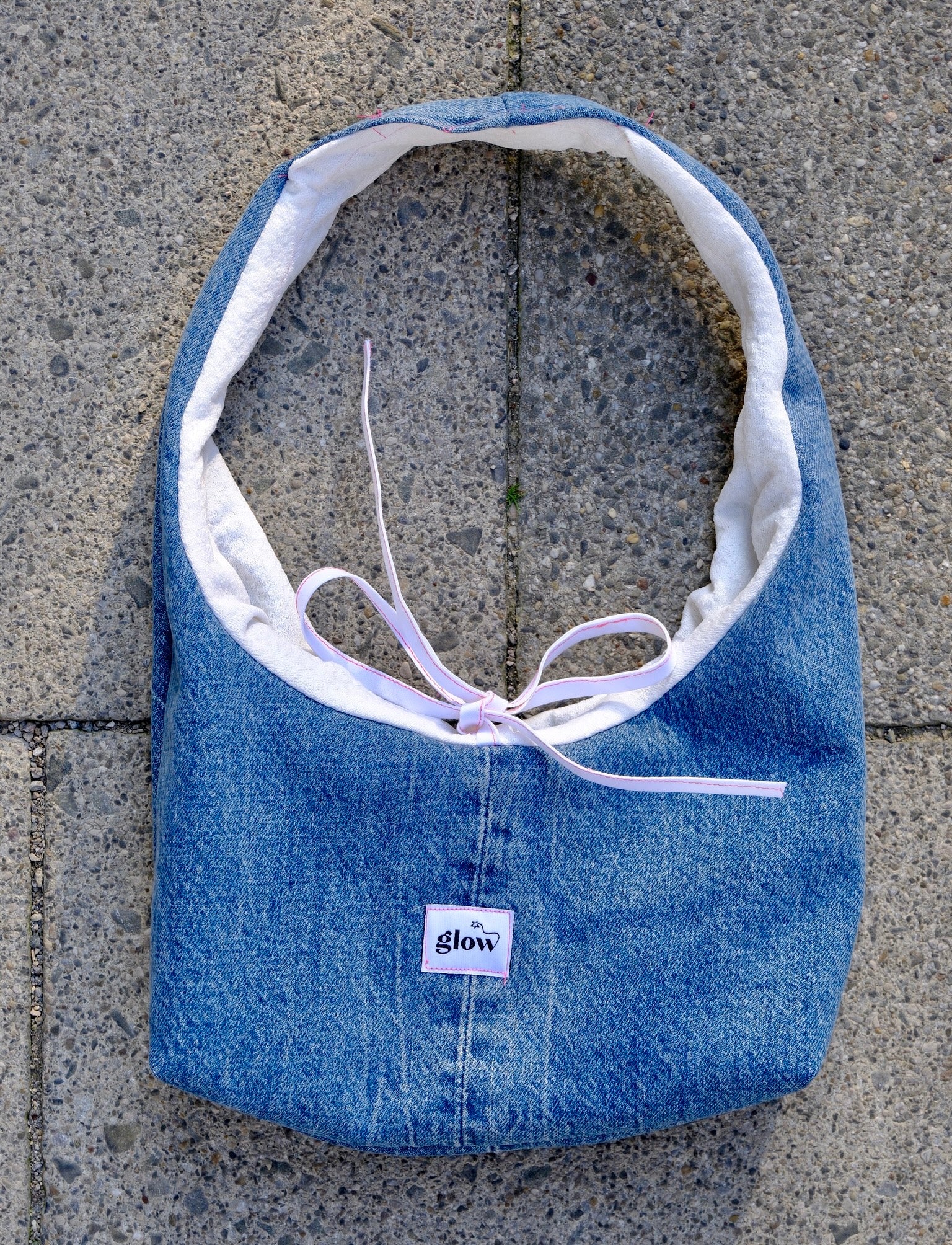 A blue denim shoulder bag with a white interior lining and a light purple tie, laid flat on gray concrete pavement. A small white label on the front reads 'glow'.