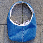 A blue denim shoulder bag with a white lining and a light purple tie, laid flat on gray stone pavers.