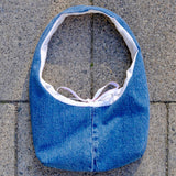 A blue denim shoulder bag with a white lining and a light purple tie, laid flat on gray stone pavers.