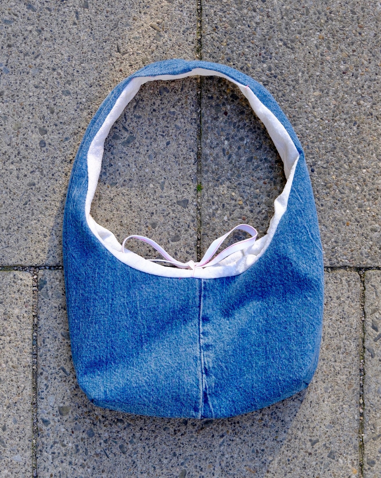 A blue denim shoulder bag with a white lining and a light purple tie, laid flat on gray stone pavers.