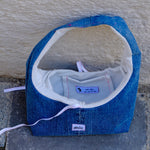 A handmade blue denim shoulder bag with a white cotton lining, resting on a gray stone surface against a textured beige stucco wall. The brand name 'blanco' is visible on a small white patch on the front.