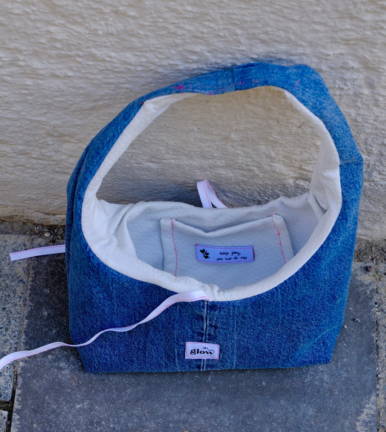 A handmade blue denim shoulder bag with a white cotton lining, resting on a gray stone surface against a textured beige stucco wall. The brand name 'blanco' is visible on a small white patch on the front.