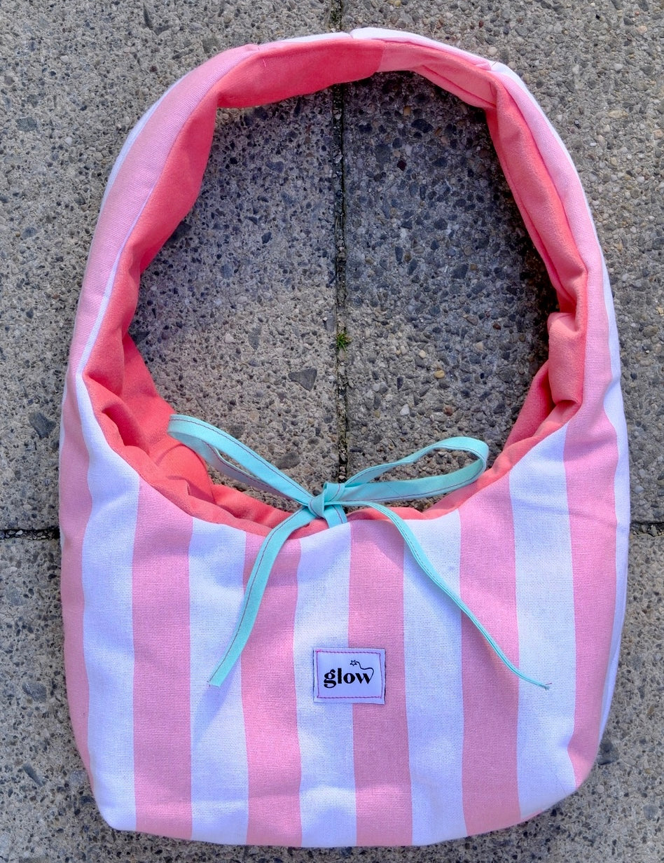 A fabric shoulder bag with vertical pink and white stripes, a coral pink lining, and a light blue ribbon tie, laid flat on a gray concrete surface. A small label on the front reads 'glow'.