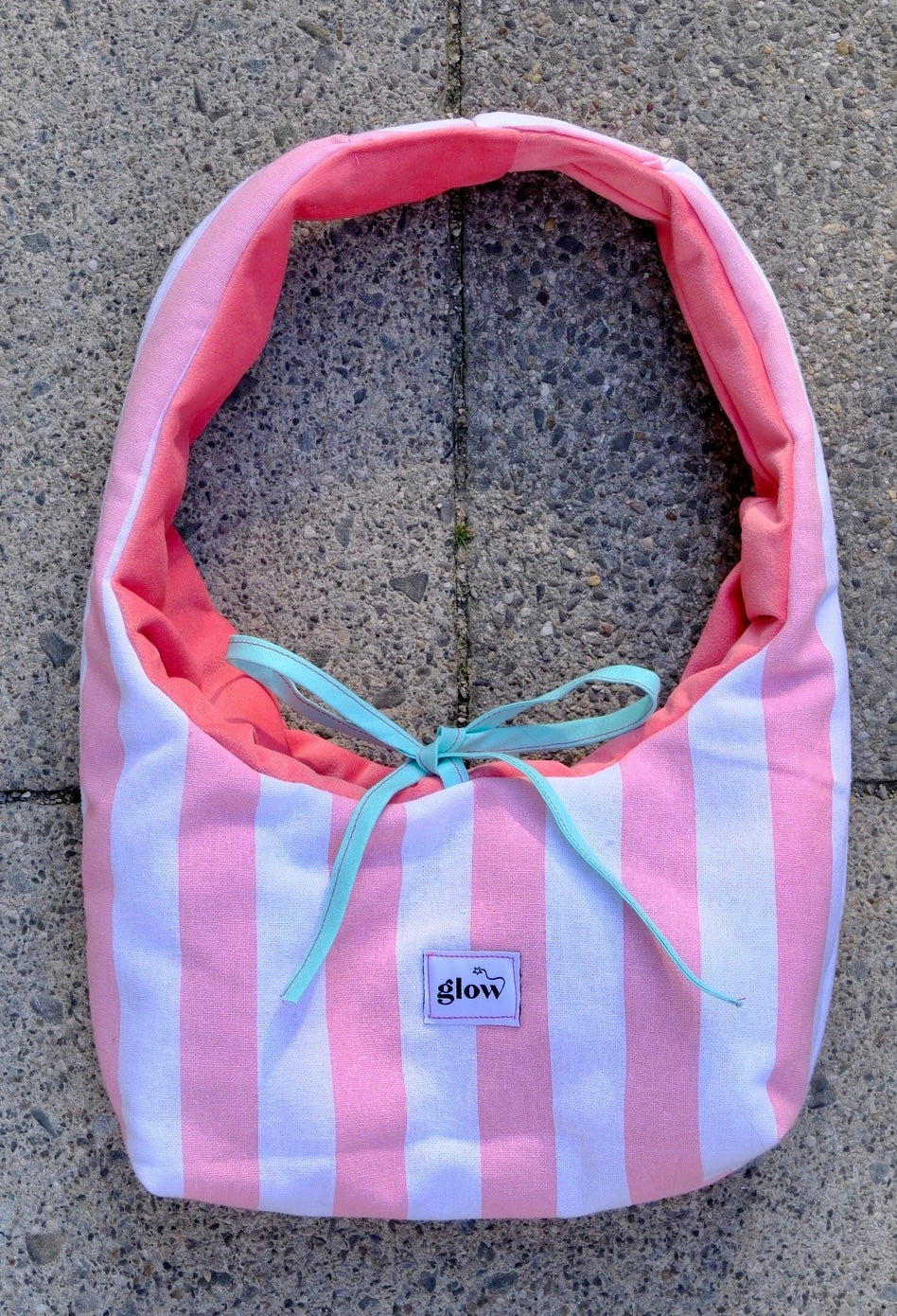 A fabric shoulder bag with vertical pink and white stripes, a coral pink lining, and a light blue ribbon tie, laid flat on a gray concrete surface. A small label on the front reads 'glow'.