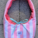 A fabric shoulder bag with vertical pink and white stripes, a coral pink lining, and a light blue ribbon tie, laid flat on a gray concrete surface. A small label on the front reads 'glow'.