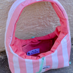 A pink and white striped dog crate with a pink interior, shown outdoors against a textured beige wall and gray pavement. A small sewn label on the front reads glow.