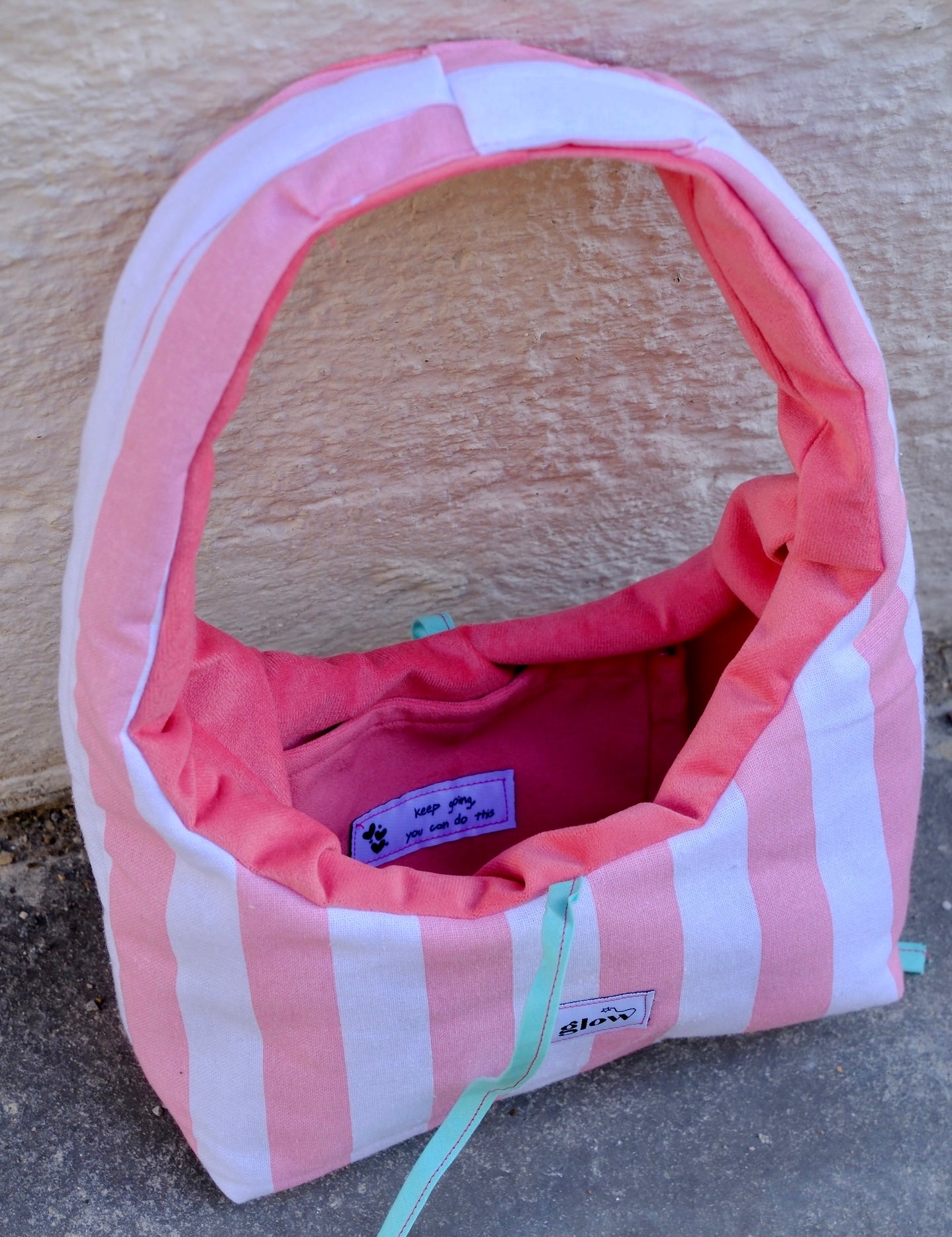 A pink and white striped dog crate with a pink interior, shown outdoors against a textured beige wall and gray pavement. A small sewn label on the front reads glow.