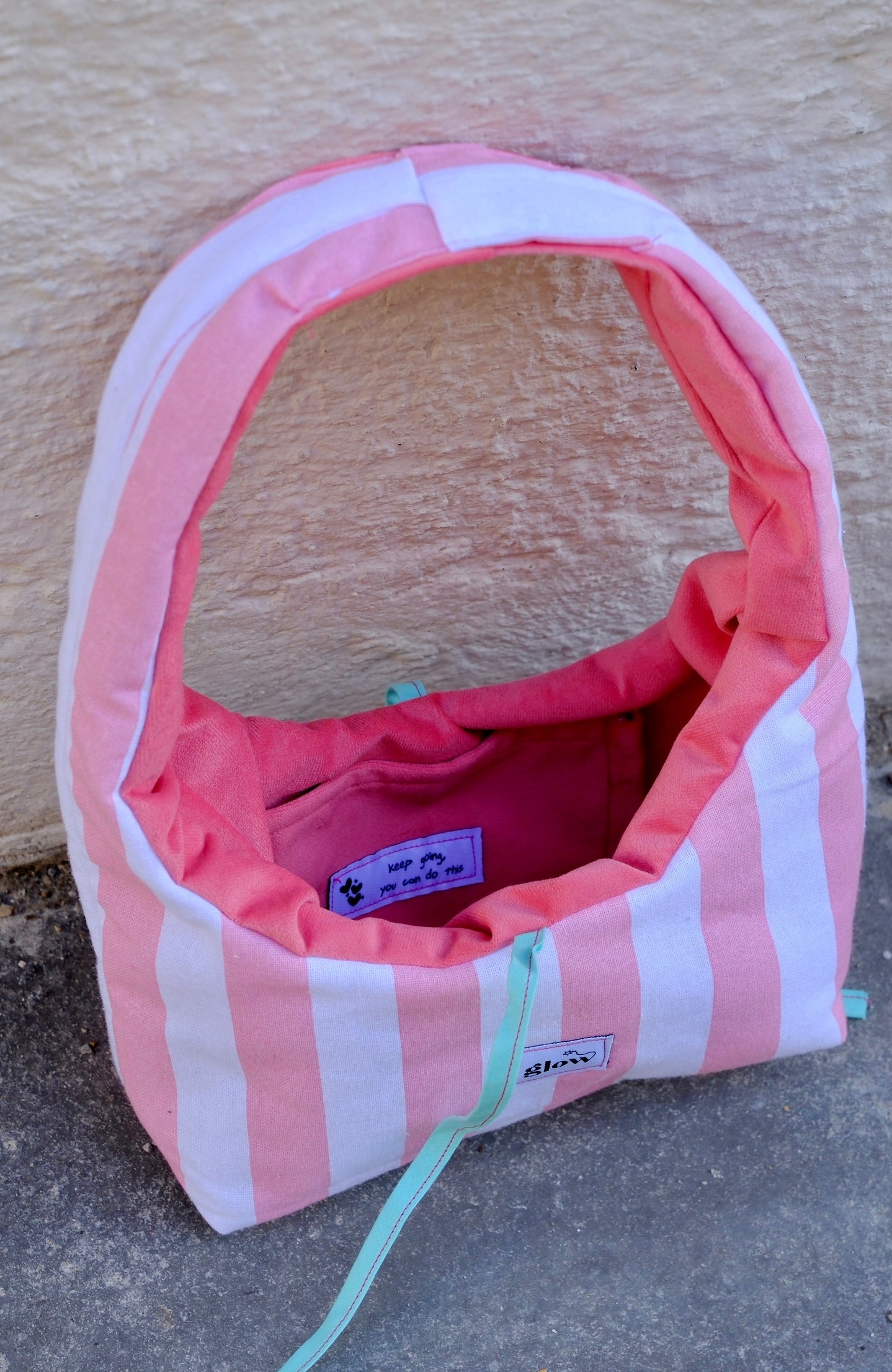 A pink and white striped dog crate with a pink interior, shown outdoors against a textured beige wall and gray pavement. A small sewn label on the front reads glow.